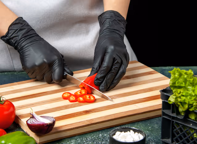 Gloved hands slice red chili peppers on a striped cutting board with nearby vegetables.