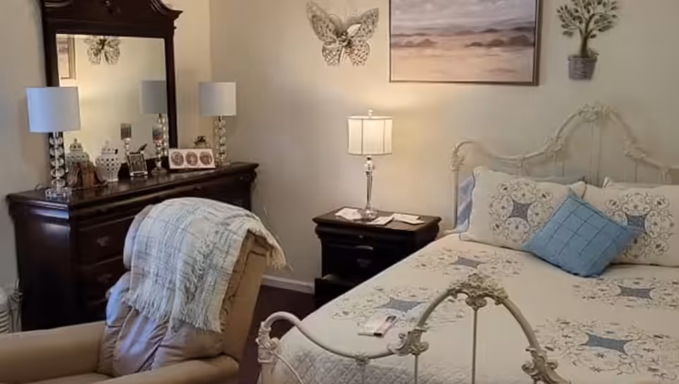 A cozy bedroom featuring a decorative metal bed frame with a quilted bedspread and multiple pillows, a wooden dresser with a large mirror, two table lamps, a comfortable armchair draped with a blanket, and wall decorations including a butterfly and a landscape painting.