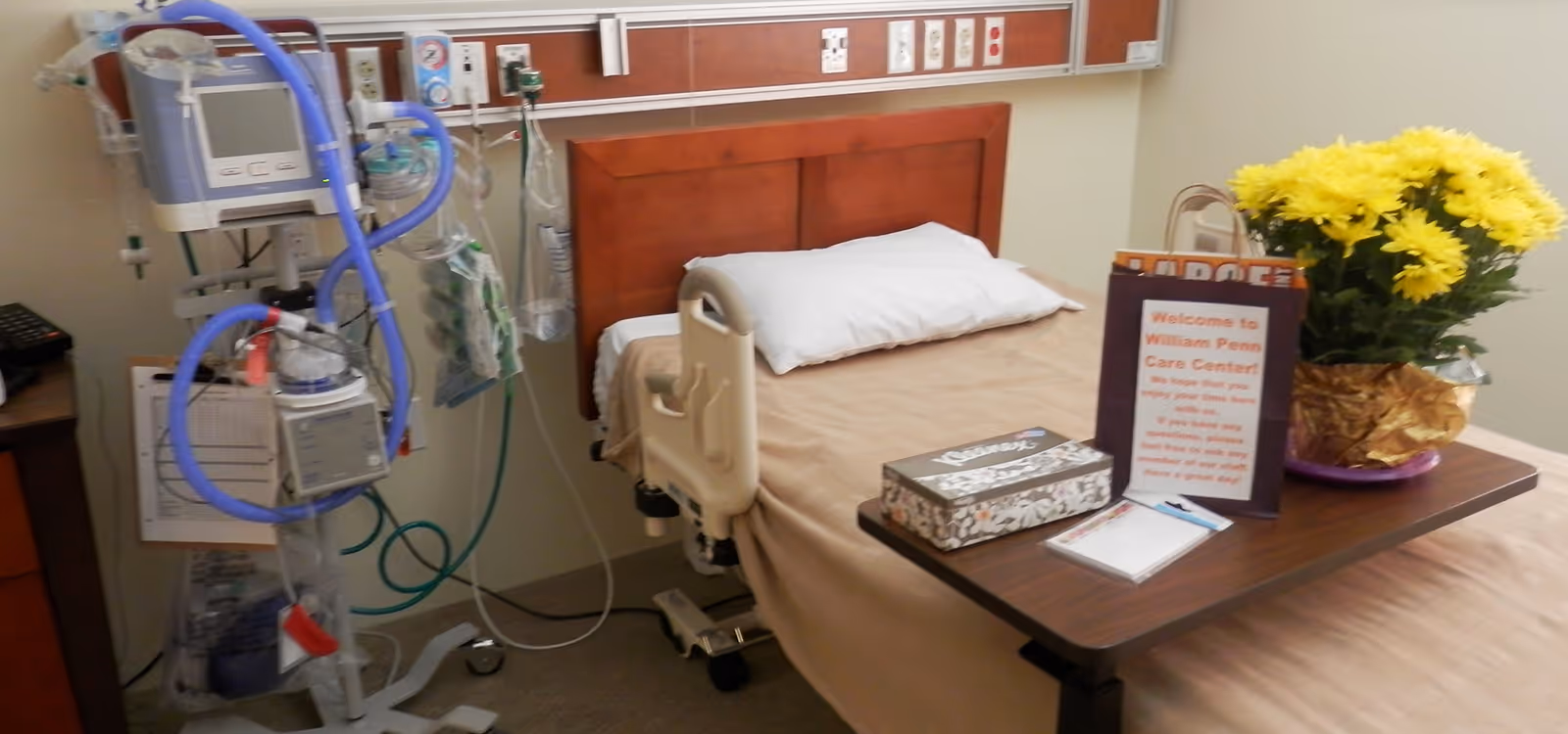 A hospital-style bed with a beige blanket and white pillow in a care center room. Next to the bed is a medical ventilator machine with tubes and monitors. A wooden bedside table holds a box of Kleenex tissues, a notepad with a pen, a welcome sign for William Penn Care Center, and a vase with yellow flowers.