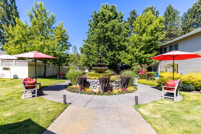 A sunny outdoor garden area with a circular stone fountain in the center, surrounded by colorful flowers and greenery. Two seating areas with chairs and red umbrellas are positioned on either side of a paved walkway. Trees and shrubs provide a lush backdrop, and a building is visible on the right side.