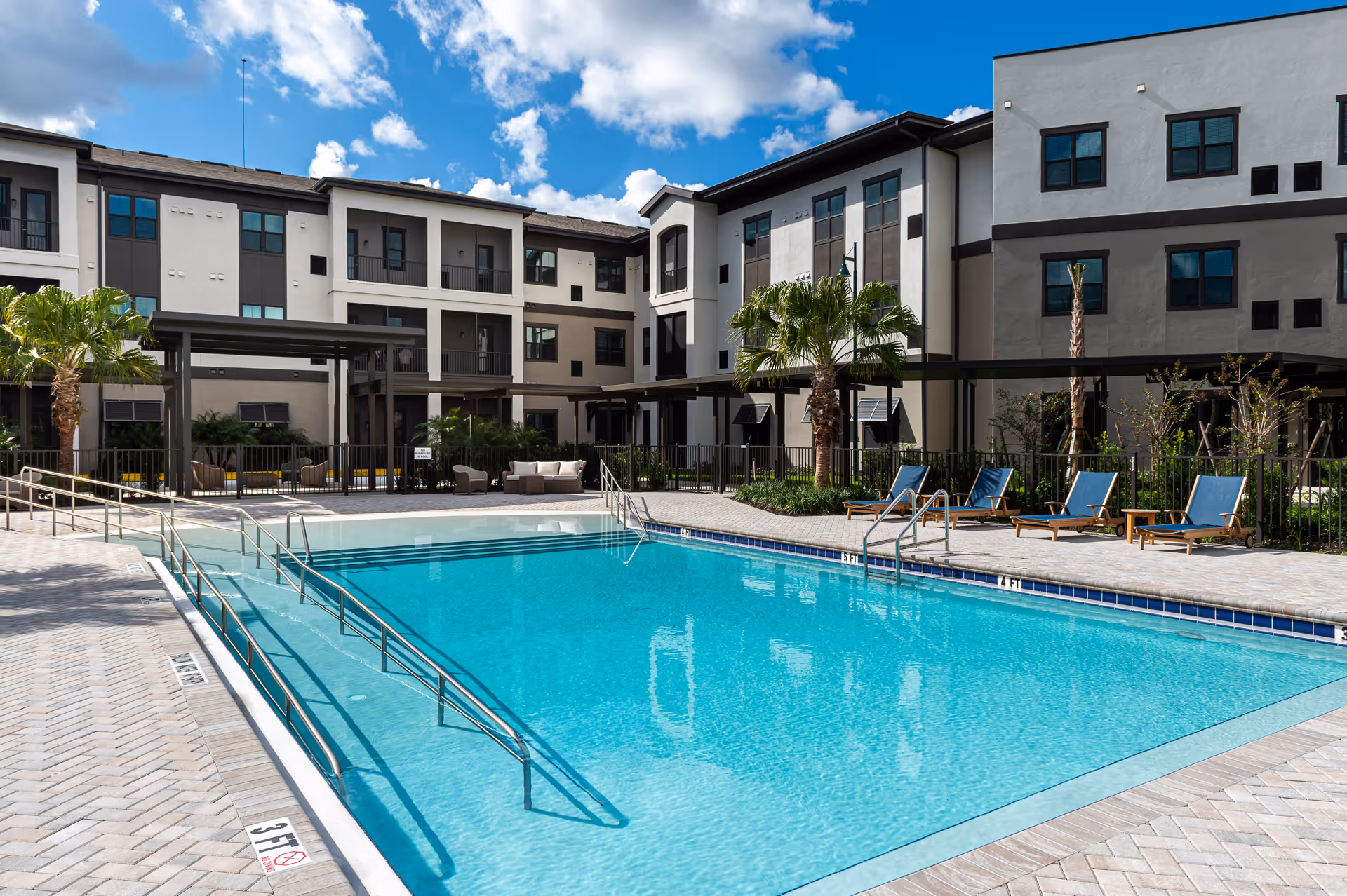 Outdoor swimming pool with lounge chairs and a multi-story residential building under a blue sky.
