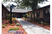 A paved walkway leading to the entrance of a single-story building with beige walls and dark trim, surrounded by landscaped garden beds with mulch, flowers, and shrubs, and a large tree providing shade near the entrance.