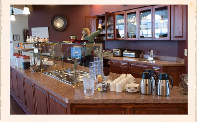 A communal serving counter and beverage station in a dining area with cups, coffee pots, glassware, and a staff member reaching into an upper cabinet.