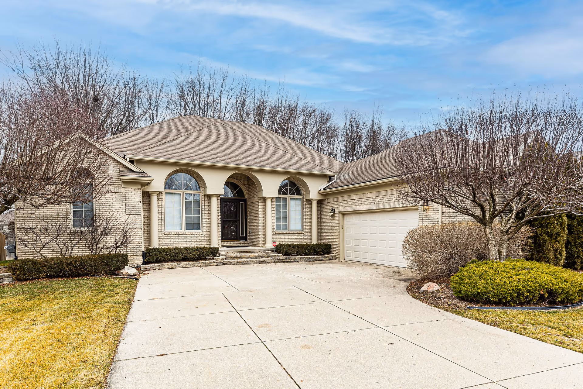 Front exterior of a single-story brick house with an arched entryway, two-car garage, driveway, and leafless trees.