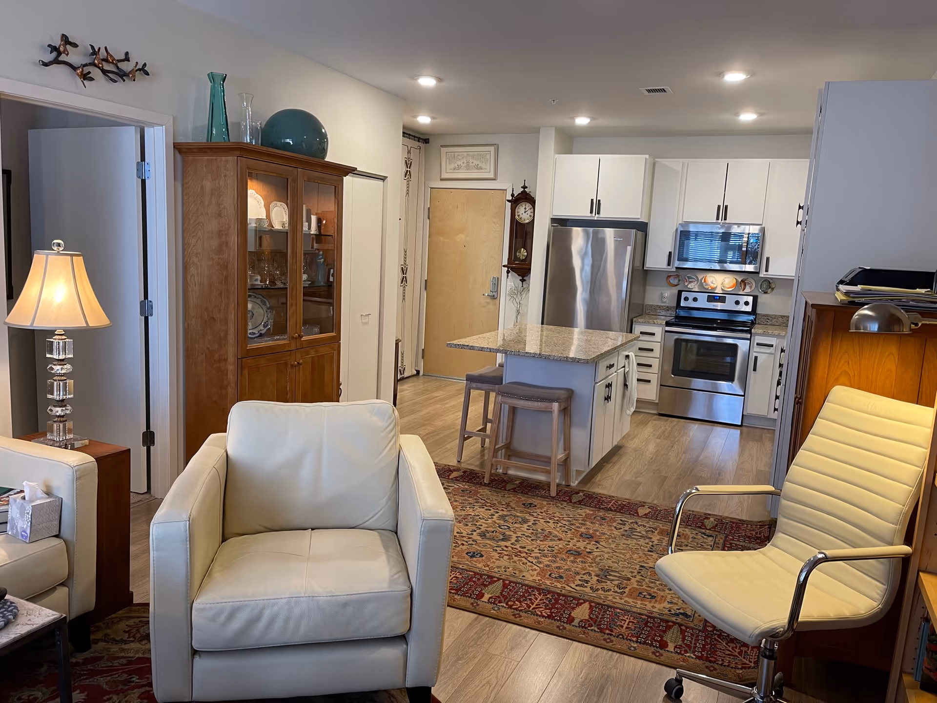 Open-plan living area with cream leather armchairs, an area rug, and a kitchen island with stainless steel appliances in the background.