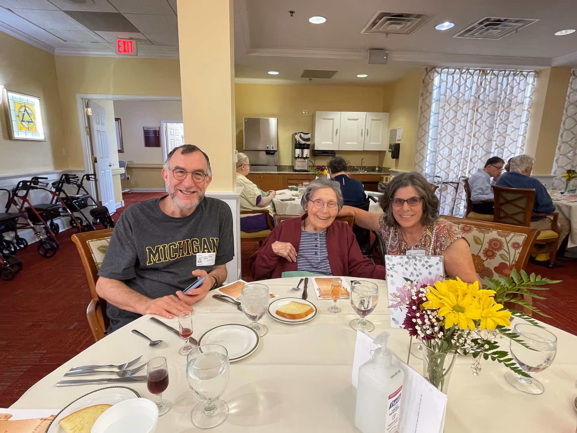 Three people smiling at a dining table set with plates, glasses, and a vase of yellow flowers in an assisted-living dining room.