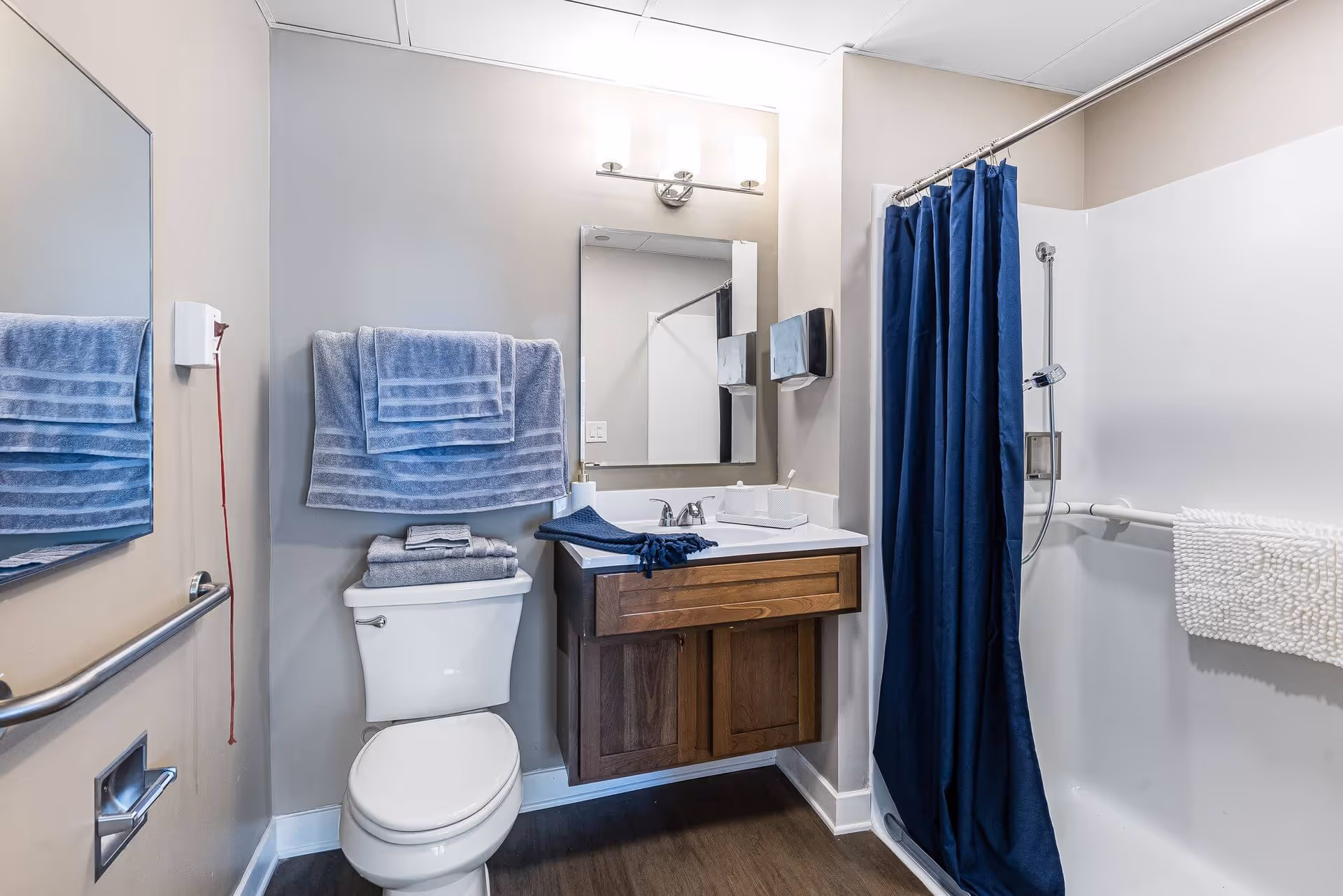 Accessible bathroom with a toilet, wooden vanity and sink, mirror, blue towels, and a shower with a navy curtain and grab bars.