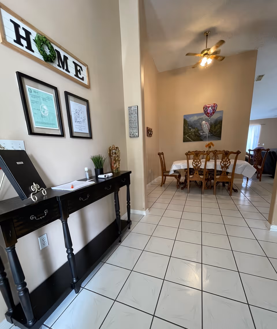 Interior view of a home care facility showing a hallway with a black console table against the left wall, decorated with framed documents and a 'HOME' sign above it. The hallway leads to a dining area with a wooden dining table and chairs, a ceiling fan with lights, a painting on the wall, and two heart-shaped balloons tied to the table.