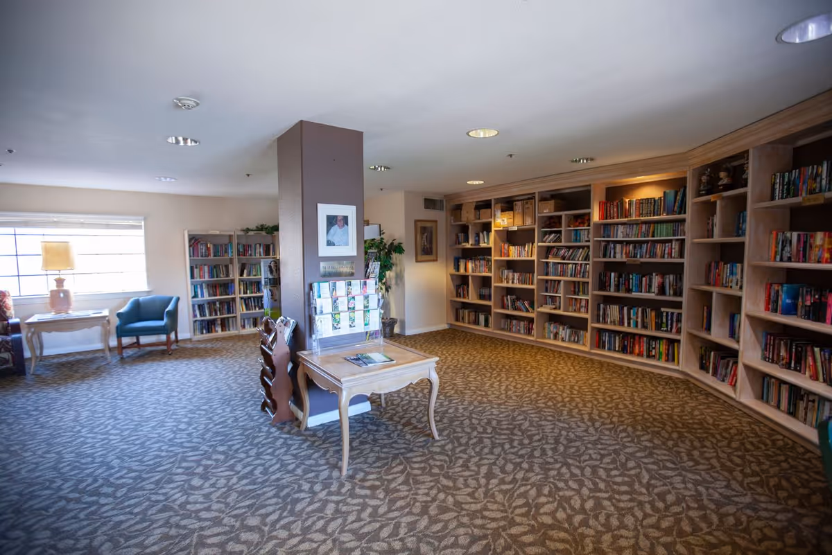 A spacious library or reading room with carpeted floor and patterned design. The room features multiple wooden bookshelves filled with books along the walls. In the center, there is a small wooden table with brochures and a magazine rack next to a brown pillar. A blue armchair and a side table with a lamp are positioned near a window letting in natural light. The room has a calm and inviting atmosphere.