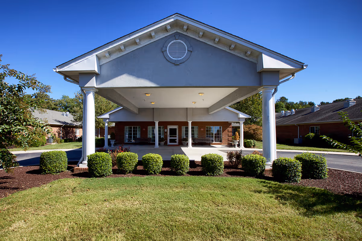 Front exterior view of a single-story brick building with a covered entrance supported by white columns, surrounded by neatly trimmed bushes and green lawn under a clear blue sky.