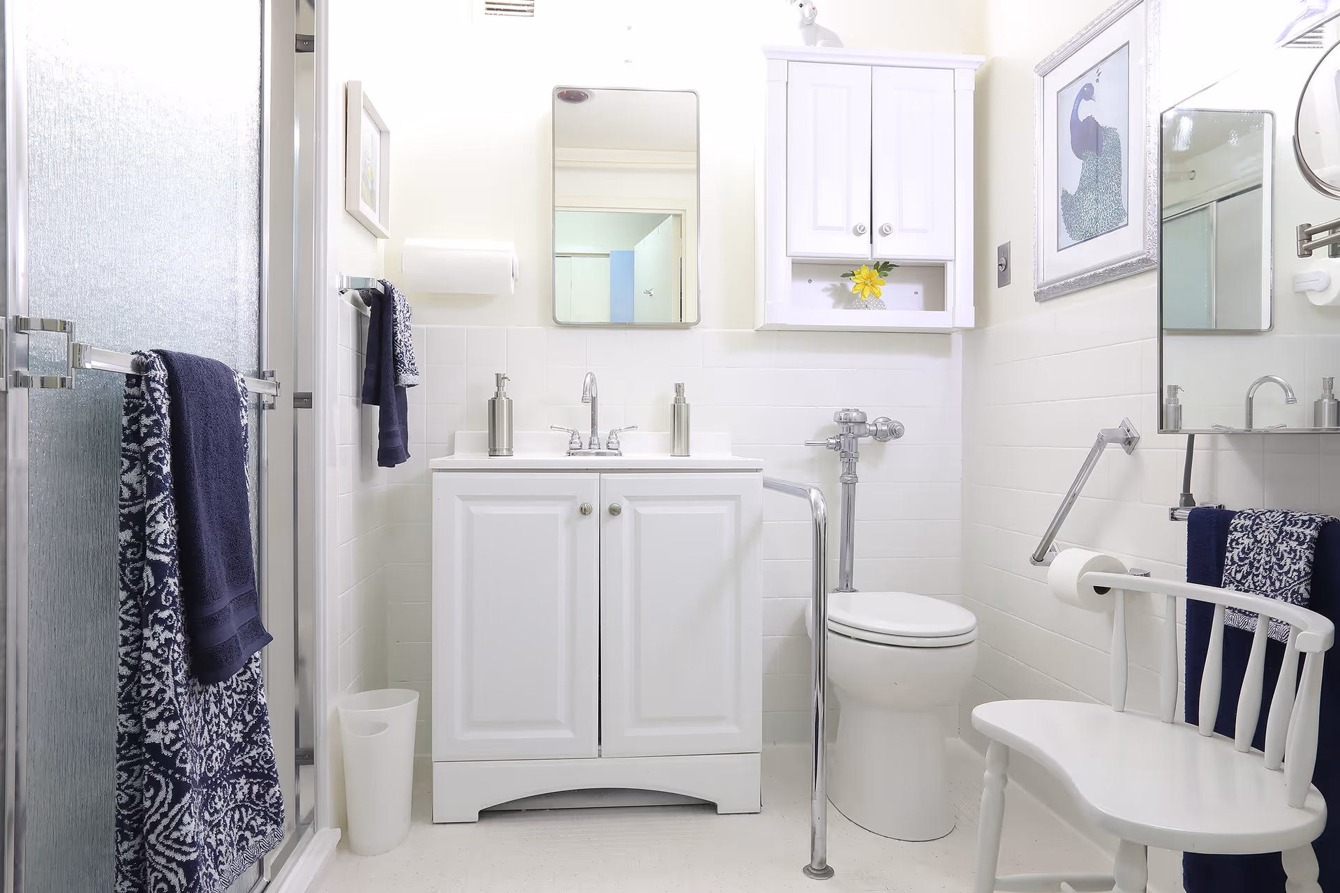 A bright and clean bathroom with white walls and fixtures. There is a shower with frosted glass on the left, a white vanity with a sink and mirror in the center, and a toilet with a safety grab bar on the right. A white chair with a blue patterned towel is positioned near the toilet. The bathroom also features a wall-mounted cabinet above the toilet and framed artwork on the wall.