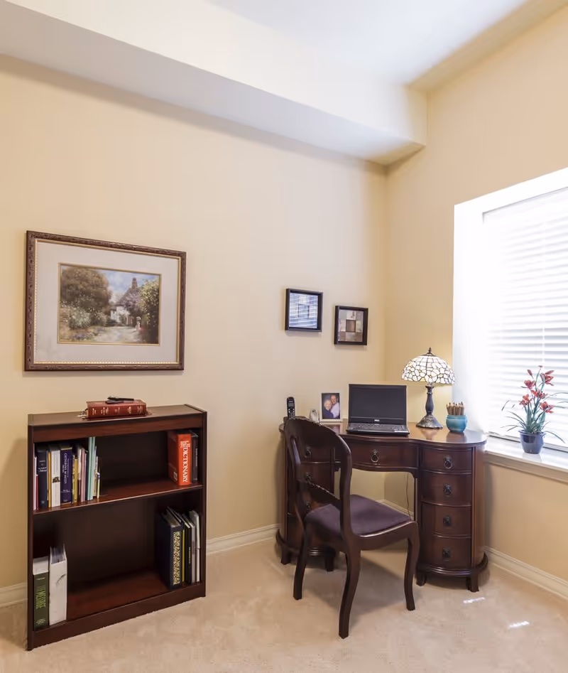 A cozy corner office space in a senior living facility with a wooden desk and chair. On the desk are a laptop, a decorative lamp, a small plant, a photo frame, and a telephone. Next to the desk is a small bookshelf with books and binders. The walls are painted beige and decorated with framed artwork. A window with white blinds allows natural light to brighten the room.