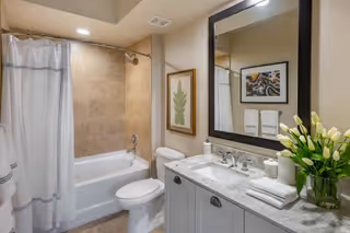 A clean and modern bathroom featuring a bathtub with a shower curtain, a toilet, a vanity with a marble countertop, a large mirror, a framed botanical print on the wall, and a vase of white tulips on the counter.