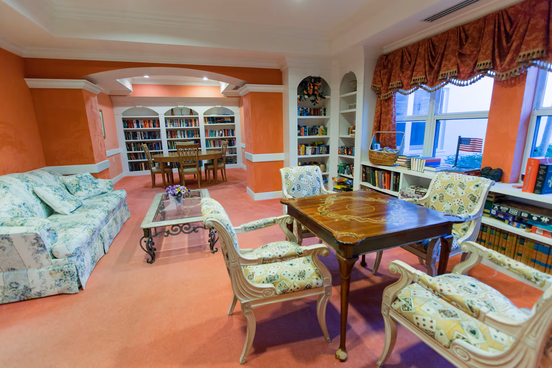 A cozy common area with coral-colored walls and carpet, featuring a floral-patterned sofa, a glass coffee table, and a wooden game table surrounded by four cushioned chairs with playing card designs. Built-in bookshelves filled with books and board games line the walls, and a window with red valances lets in natural light.