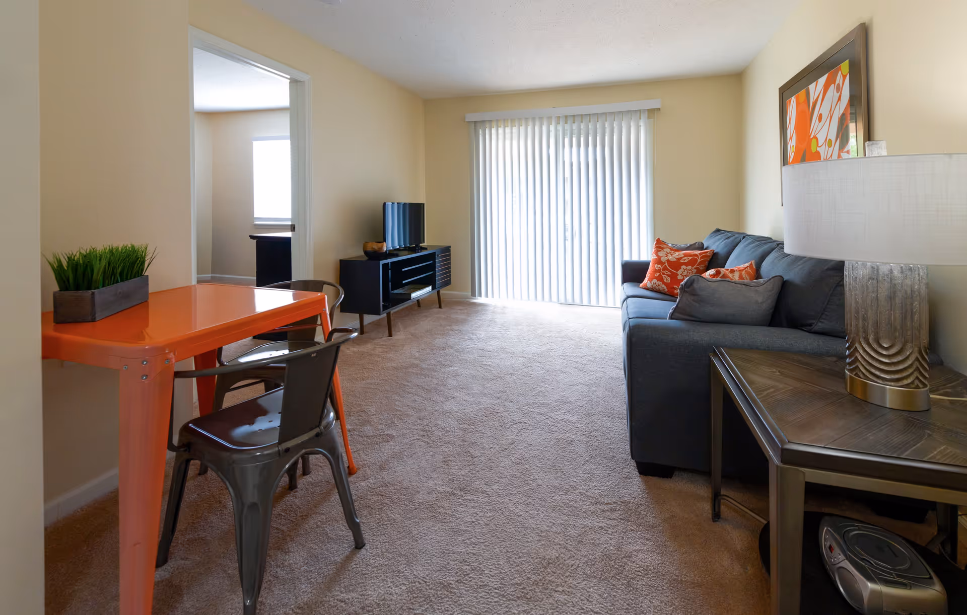 A living room with beige walls and carpet, featuring a gray sofa with orange and gray pillows, a side table with a decorative lamp, an orange table with two metal chairs, a TV on a black stand, and vertical blinds covering a sliding glass door.