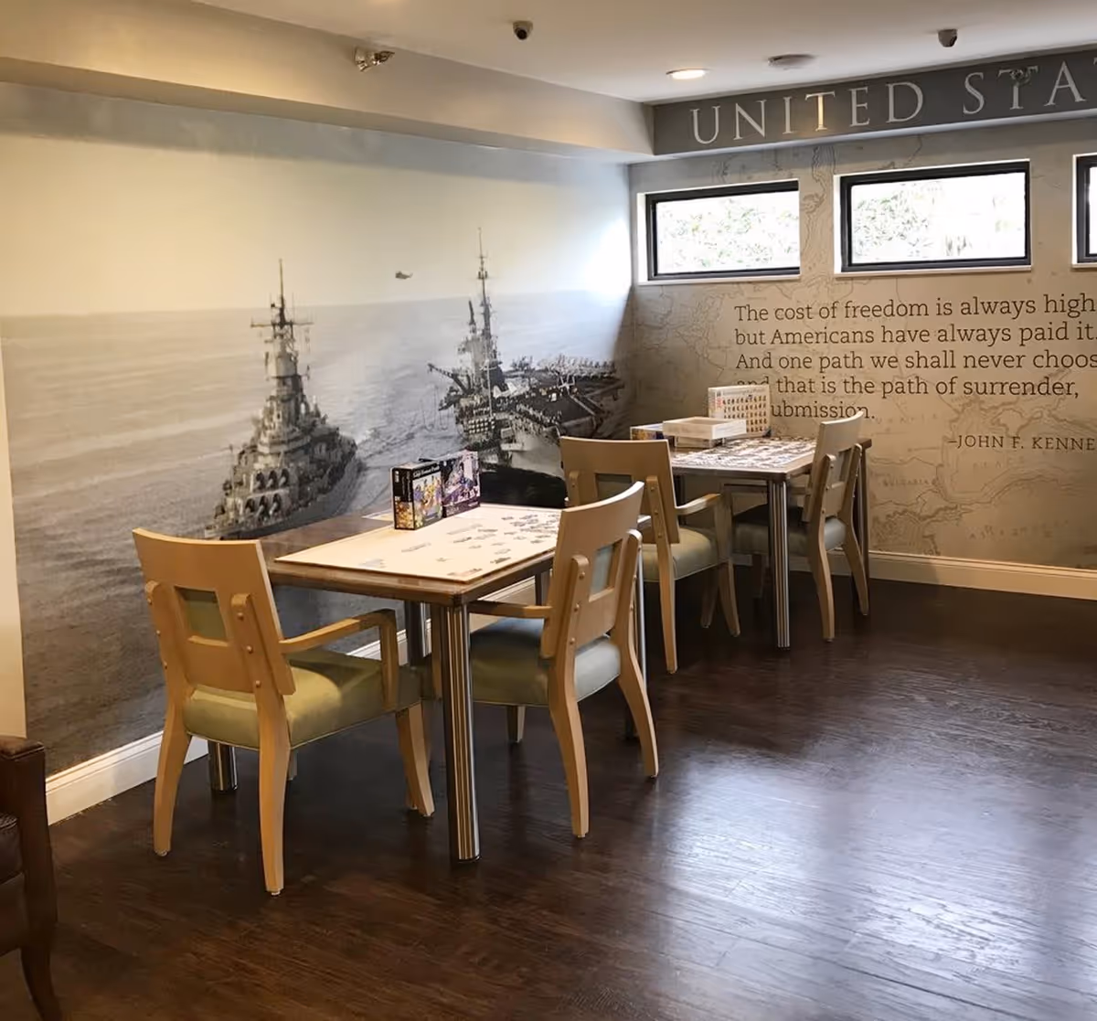 A room with two tables and several chairs arranged around them, with puzzles and puzzle boxes on the tables. The walls feature a large mural of battleships on the ocean and a quote from John F. Kennedy about the cost of freedom. Three small windows are above the quote, letting in natural light.