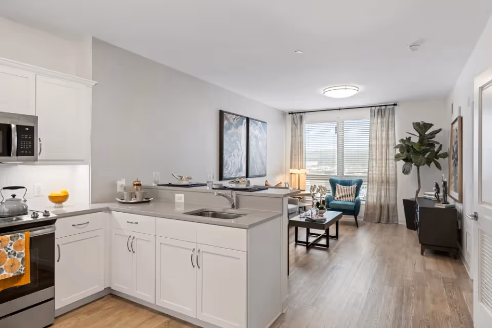 Open-plan kitchen and living area with white cabinets, a sink island, wooden floors, and a windowed seating area with a teal armchair.
