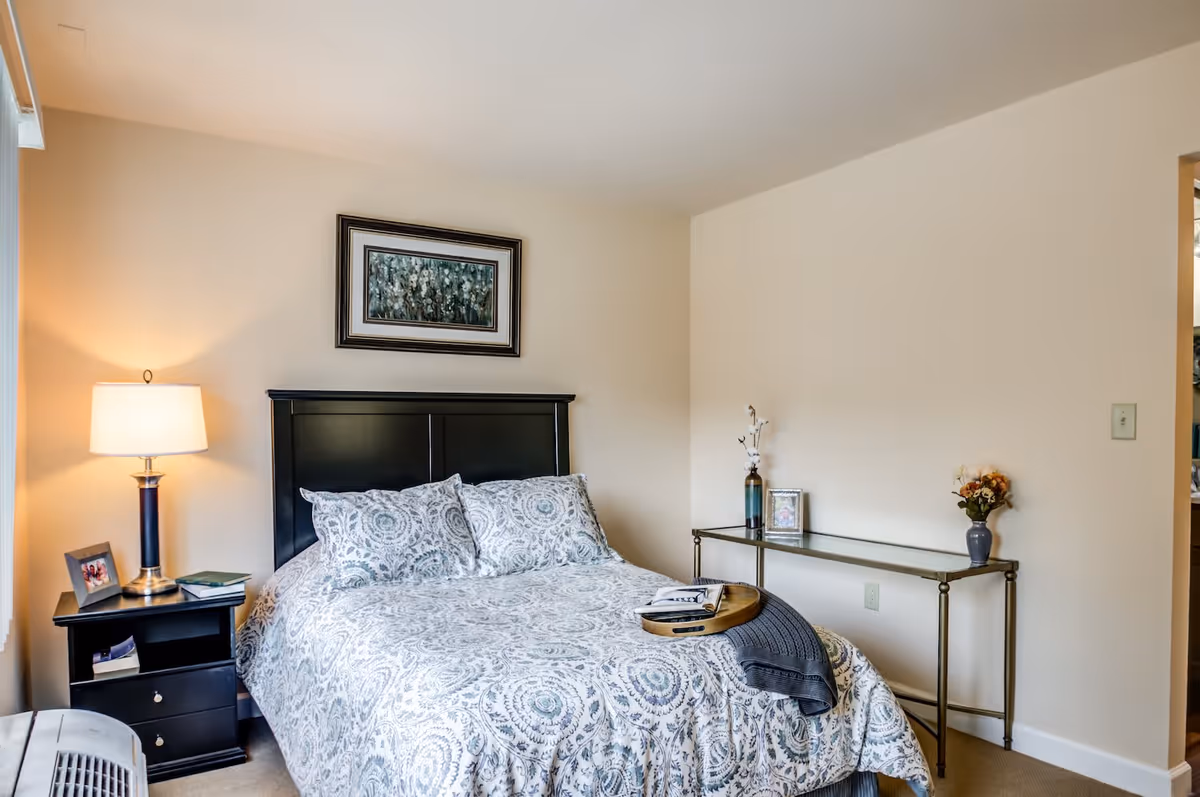A neatly made bed with a patterned bedspread and two matching pillows in a bedroom. To the left of the bed is a black nightstand with a lamp, a framed photo, and some books. Above the bed hangs a framed piece of artwork. On the right side of the room is a narrow glass console table with decorative items including a vase with flowers and a small framed picture. The walls are painted beige and the room is softly lit.