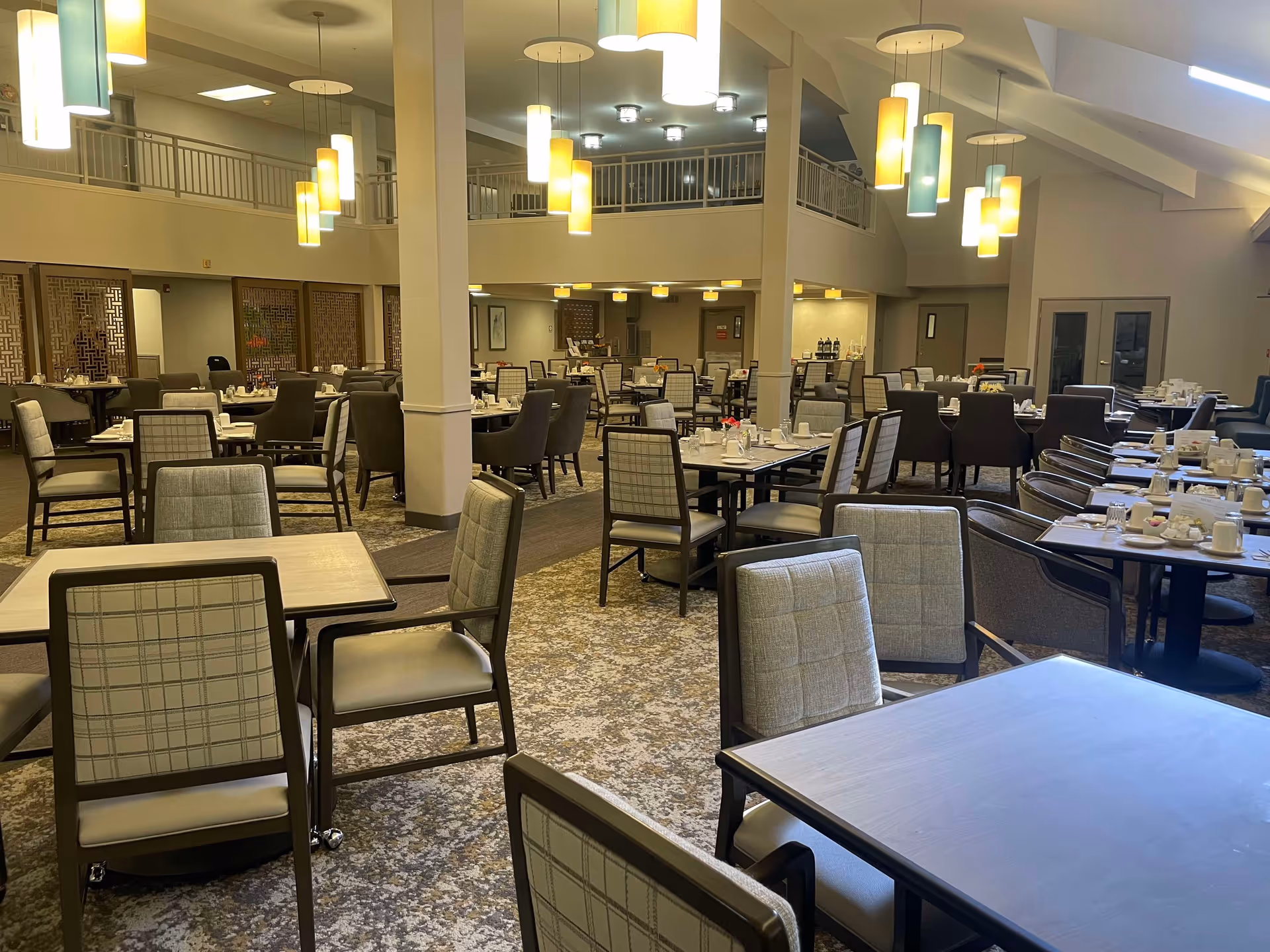 Spacious dining room with multiple tables and chairs arranged neatly. The room features modern hanging pendant lights, a patterned carpet, and a second-floor balcony overlooking the area. Tables are set with cups, plates, and utensils, ready for meal service.