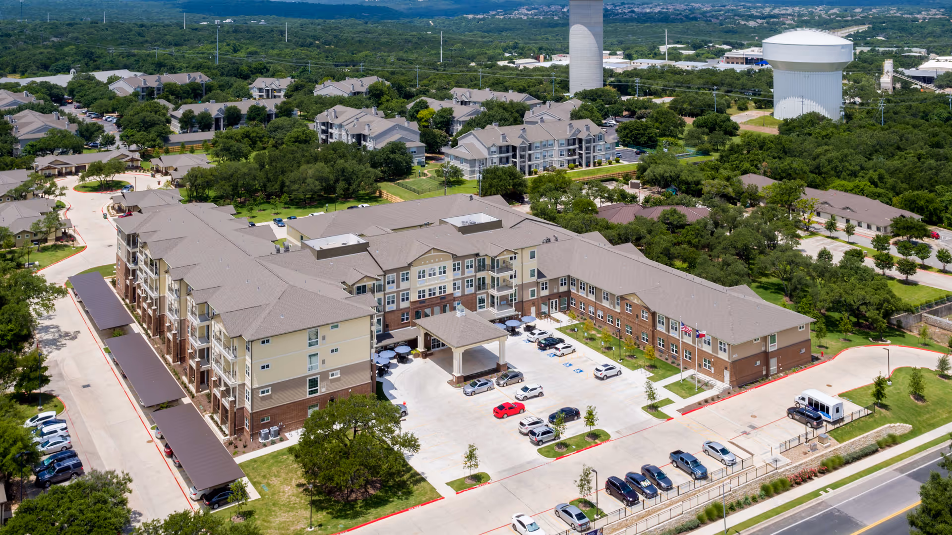 Aerial view of The Enclave at Cedar Park Senior Living facility showing a large multi-story building with a covered entrance, surrounded by parking lots and greenery. The facility is located in a suburban area with other residential buildings and a water tower visible in the background.