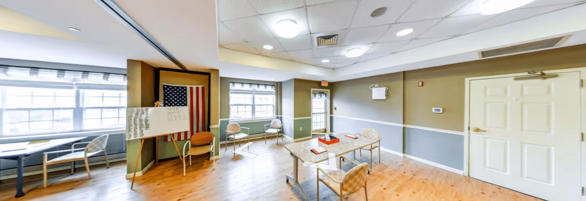 A bright, spacious room with large windows letting in natural light. The room has wooden flooring and beige and gray walls with white trim. There are several chairs arranged around a rectangular table with some books and a red box on it. An American flag is displayed on a stand next to a whiteboard with writing on it. The ceiling has recessed lighting and a white door is visible on the right side of the room.