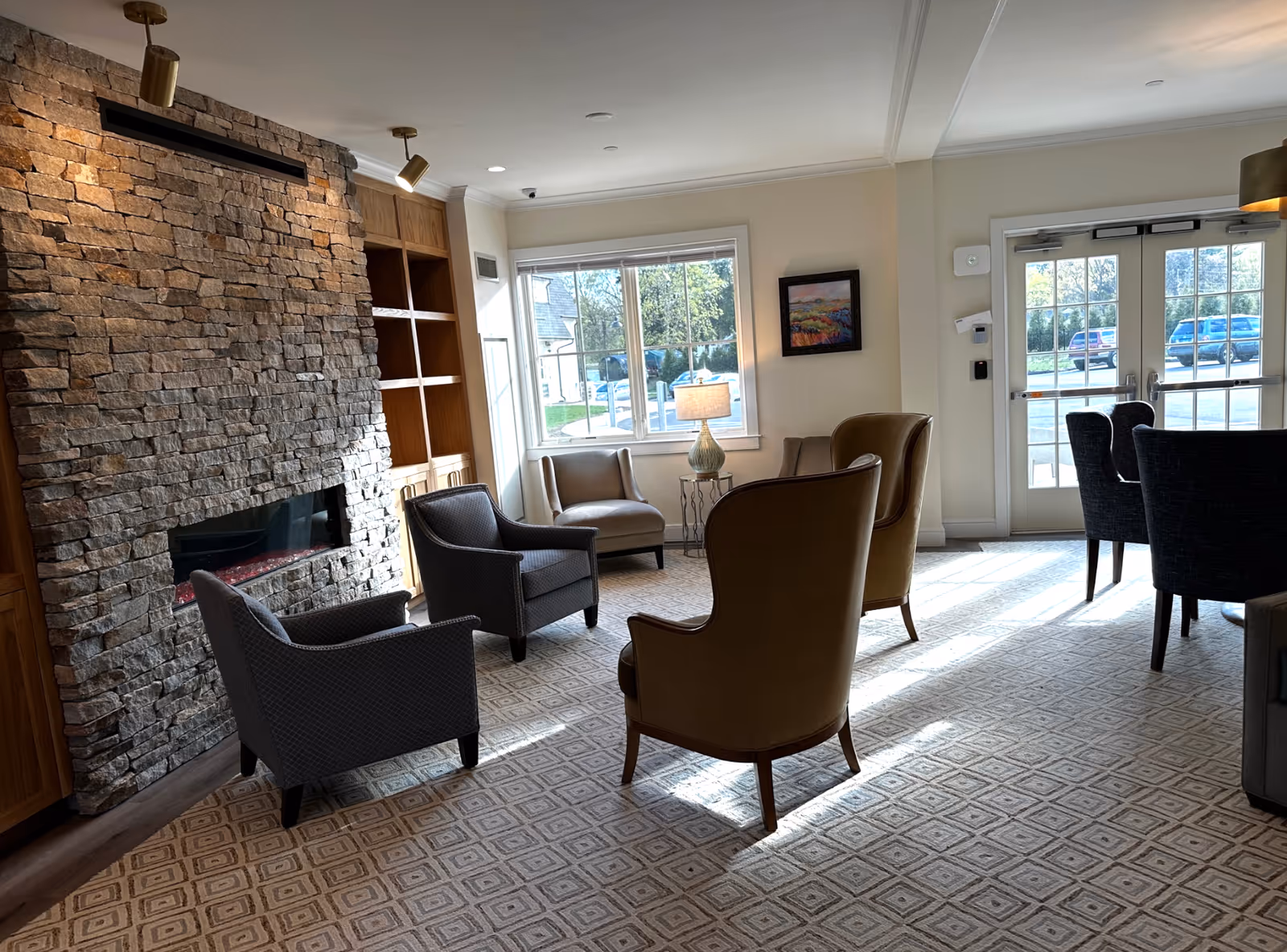 Sunlit lounge with armchairs arranged around a stone fireplace, a lamp by the window, and glass entry doors.