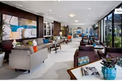 Spacious and modern living room area in a senior living facility with multiple seating arrangements including sofas and armchairs. Large windows on the right side allow natural light to fill the room. The room features contemporary decor with colorful cushions, plants, and artwork on the walls.