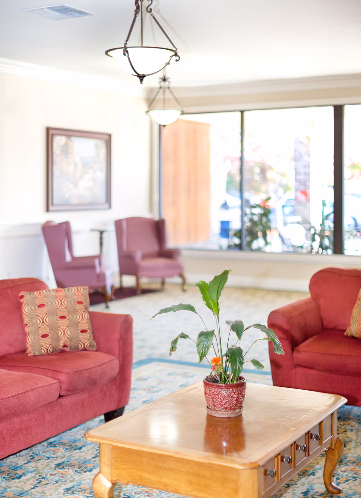 A bright and cozy living room area with two red upholstered sofas, each with patterned cushions, and two matching armchairs in the background. A wooden coffee table with a potted green plant sits in the center on a patterned carpet. Large windows allow natural light to fill the room, and two hanging light fixtures are visible on the ceiling.