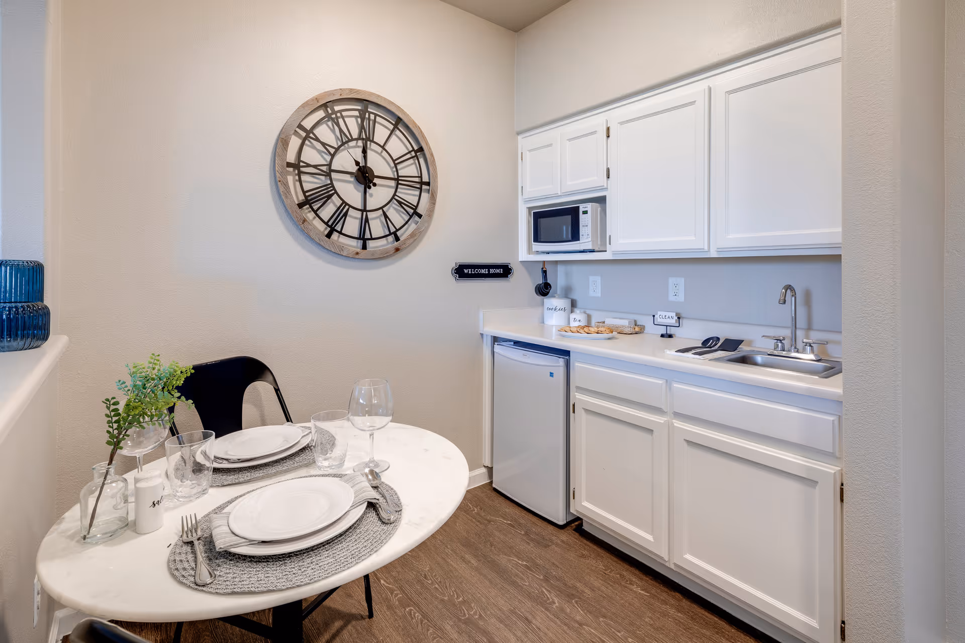 Small kitchenette and dining nook with white cabinets, a mini fridge and sink, a round table set for two, and a large wall clock.