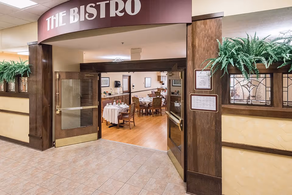 Entrance to a dining area named The Bistro in a senior living facility, featuring double wooden doors with glass panels, potted green plants on wooden ledges, and tables set with white tablecloths and chairs inside.