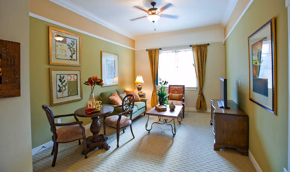 A cozy living room with green walls and beige carpet. The room features a green sofa with decorative pillows, a wooden coffee table with plants and books, a small round table with two chairs, framed artwork on the walls, a window with gold curtains, a ceiling fan with light, and a wooden TV stand with a flat-screen TV.
