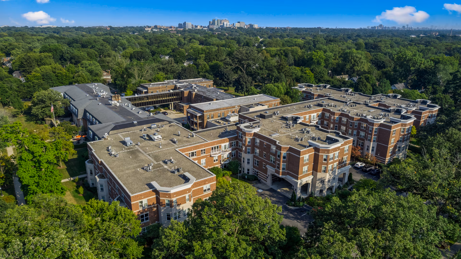 Aerial view of Three Crowns Park senior living facility surrounded by dense green trees with a city skyline in the distant background under a blue sky with scattered clouds.
