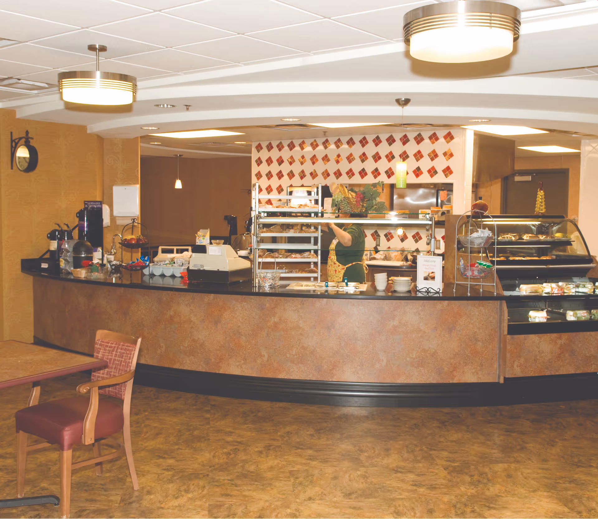 Interior view of a dining area with a curved counter displaying food items and a person behind the counter wearing an apron. The background features a wall with a red and white diamond pattern, and there are tables and chairs in the foreground.