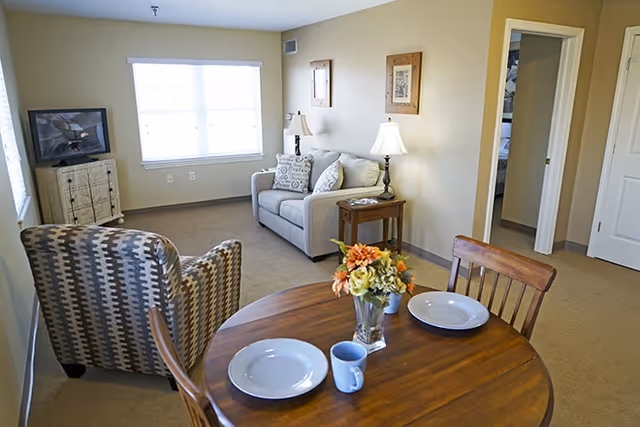 Open living area with a round dining table set for two in the foreground, an upholstered armchair and loveseat, a TV on a stand, and a window in the background.