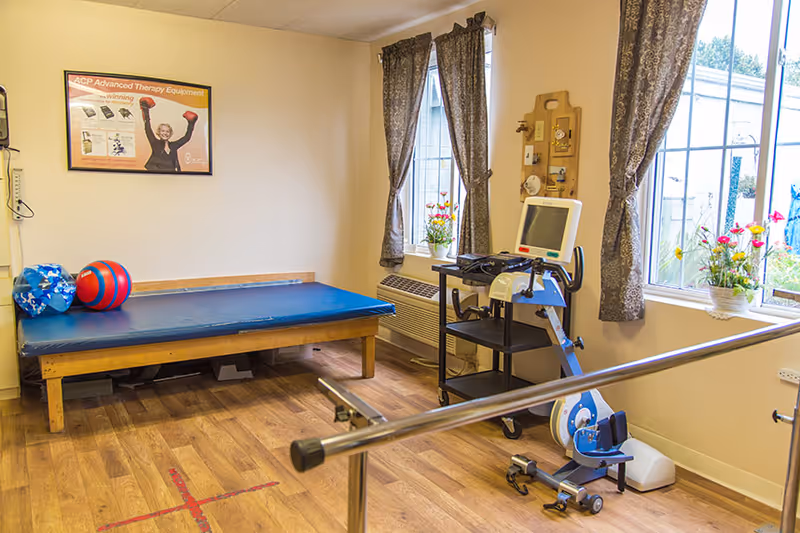 Small rehabilitation room with a treatment table, exercise equipment, parallel bars, and windows with curtains.