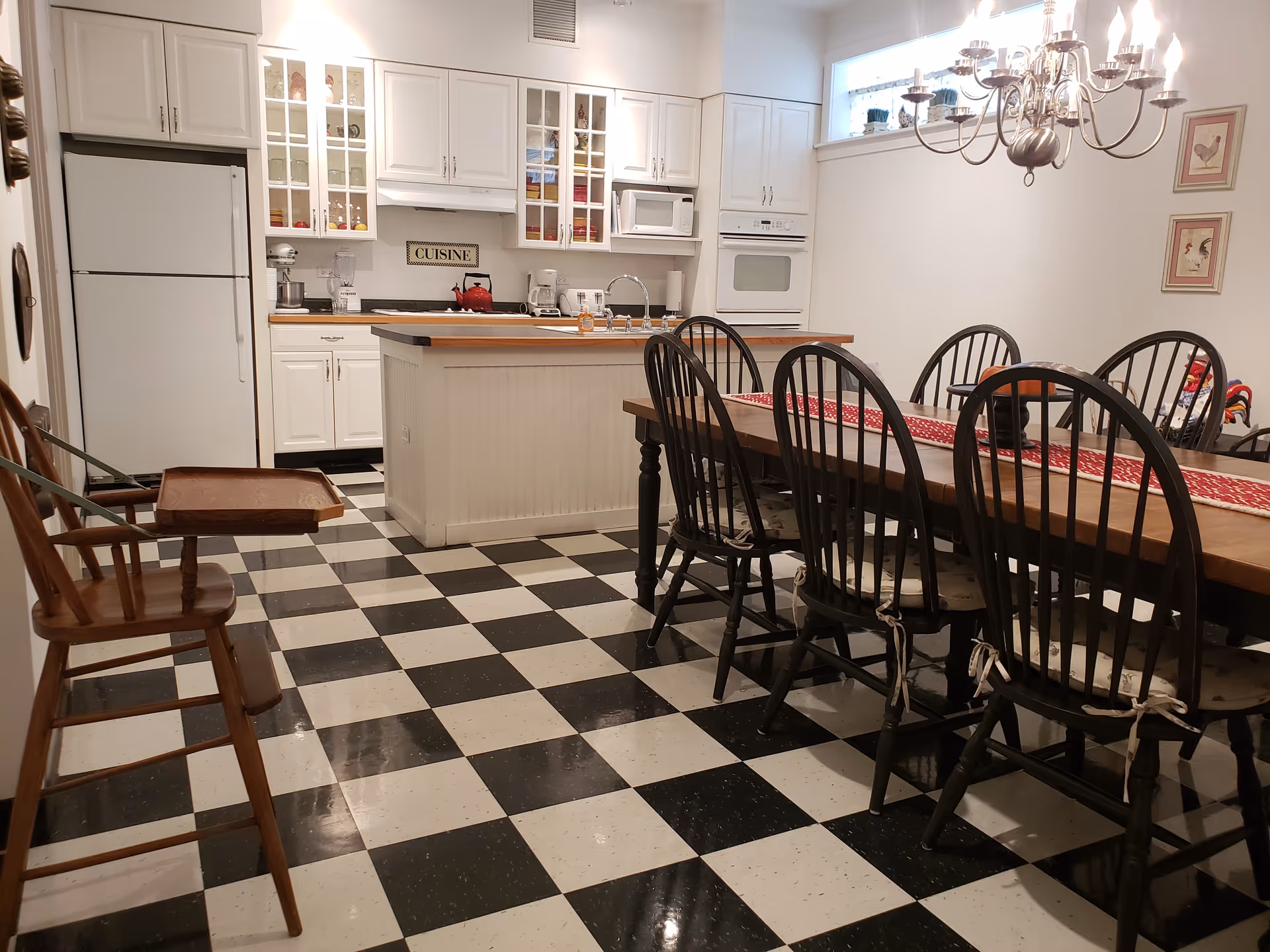 A kitchen and dining area with black and white checkered flooring. The kitchen features white cabinets, a refrigerator, a microwave, an oven, and a kitchen island with a wooden countertop and a sink. The dining area has a long wooden table with six black chairs, each with a cushion tied to the seat. A chandelier hangs above the dining table, and there are framed pictures on the wall.