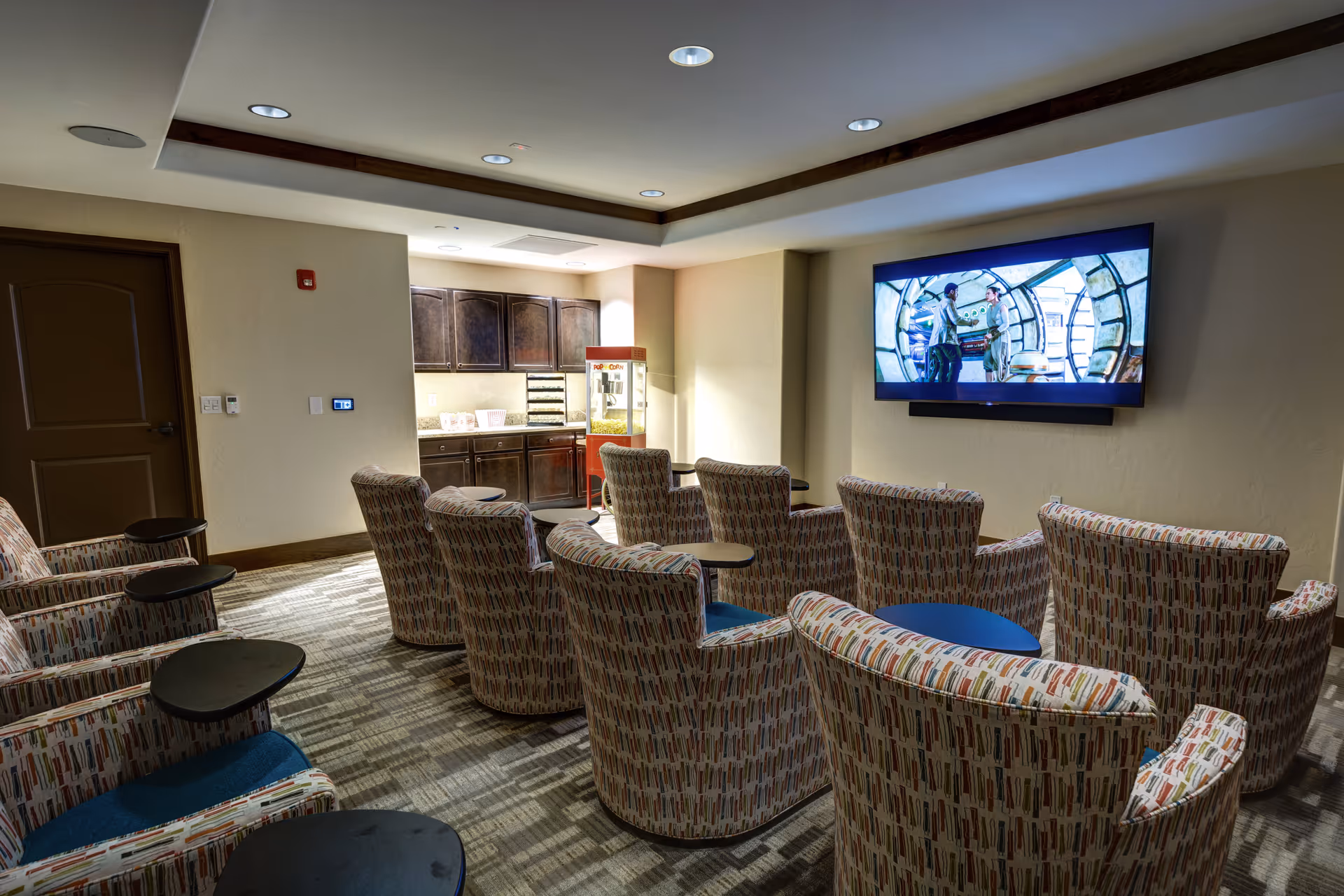 A small theater-style room with multiple patterned armchairs arranged in rows facing a large flat-screen TV mounted on the wall. Behind the chairs, there is a kitchenette area with dark wood cabinets and a popcorn machine. The room has recessed lighting and a carpeted floor.