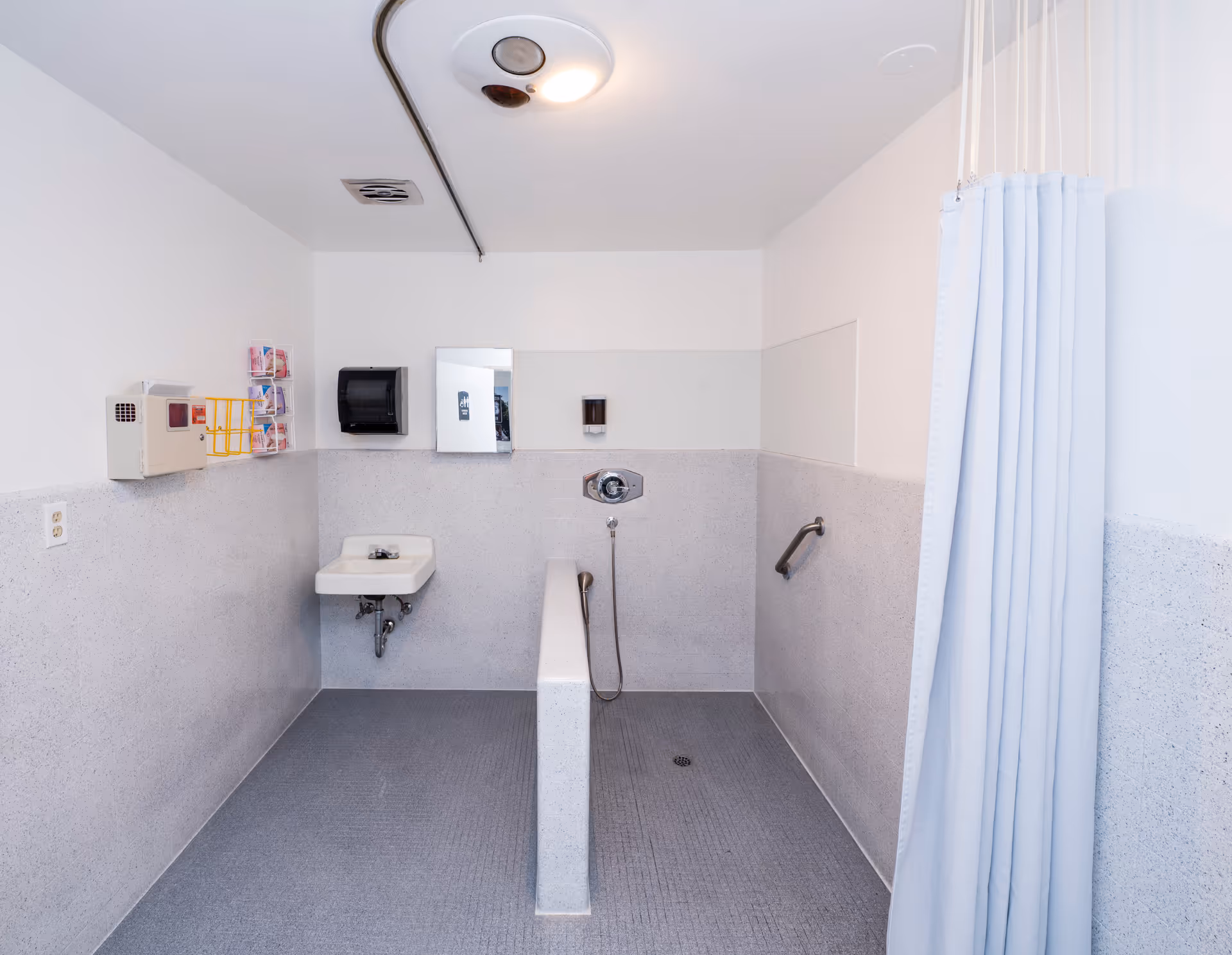 A clean, accessible shower room with a wall-mounted sink, a mirror, a soap dispenser, a hand-held shower head, grab bars, and a privacy curtain on the right side.