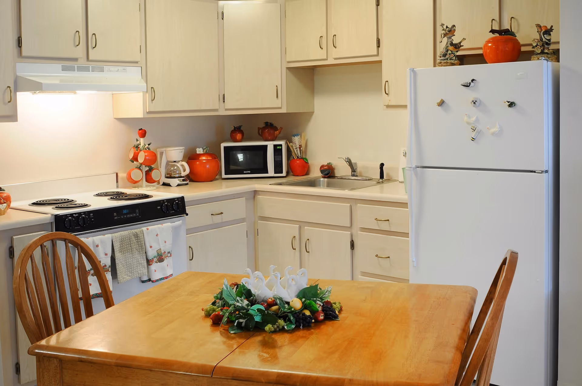 A kitchen with light wood cabinets, a white refrigerator decorated with bird magnets, a microwave, a coffee maker, and an electric stove with a white range hood. A wooden dining table with two chairs is in the foreground, featuring a decorative centerpiece with white swan figurines and artificial fruit. The kitchen has various red and orange decorative items placed on the countertops and refrigerator.