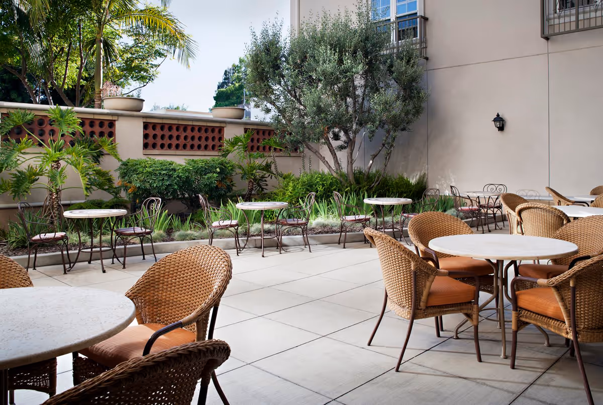 Outdoor patio area with multiple round tables and wicker chairs with orange cushions, surrounded by greenery including palm trees and bushes, adjacent to a beige building wall.