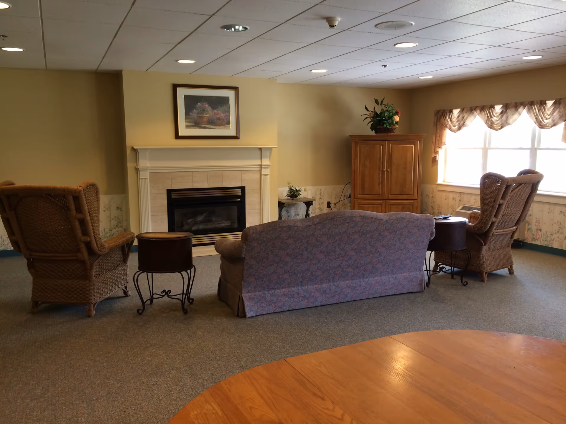A cozy living room area with a floral-patterned sofa facing a fireplace. Two wicker armchairs are positioned on either side of the sofa, each with a small round side table next to it. A wooden cabinet with a plant on top is against the wall near a window with sheer curtains. The room has beige walls, a carpeted floor, and recessed ceiling lights.