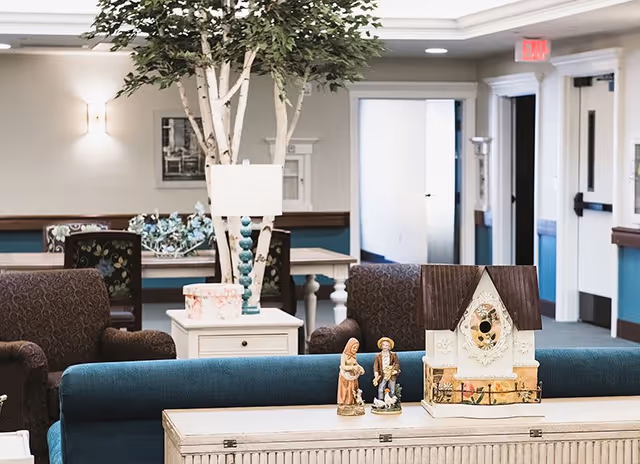 A cozy common area in a senior living facility featuring comfortable armchairs, a blue sofa, a white side table with a lamp, decorative figurines, and a birdhouse on a wooden table. There is a large indoor tree and a hallway with open doors in the background.
