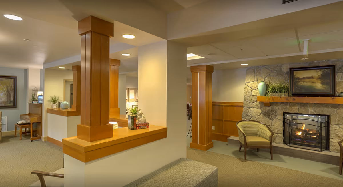 Interior view of a senior living facility common area featuring wooden pillars, a stone fireplace with a fire burning, a framed painting above the fireplace, a green vase and plants on the mantel, and comfortable seating including an armchair. The area is warmly lit with recessed ceiling lights and has carpeted flooring.