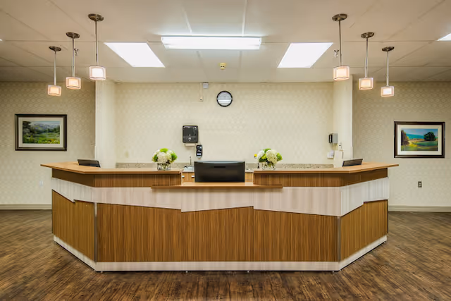Reception desk area in a senior living facility with a wooden counter, two flower arrangements, a computer monitor, and a clock on the wall behind. The room has patterned wallpaper, two framed landscape paintings, pendant lights hanging from the ceiling, and wood flooring.