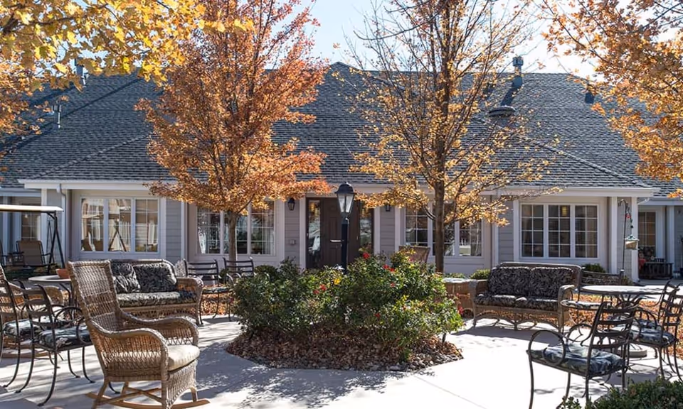 Courtyard patio with wicker and metal seating arranged around a central planter and trees in front of a low residential building.