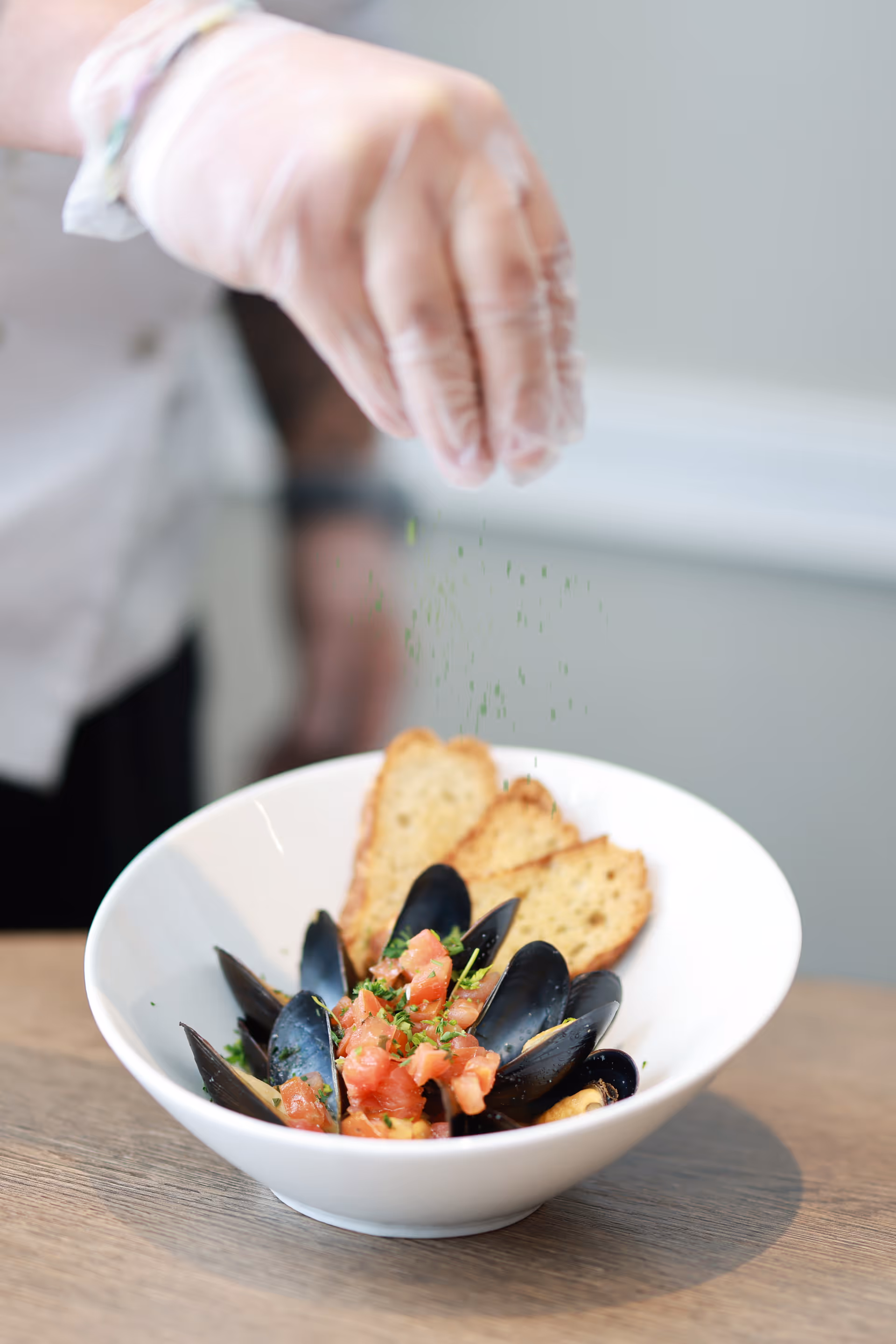 A gloved hand sprinkling herbs over a bowl of mussels topped with diced tomatoes and toasted bread.