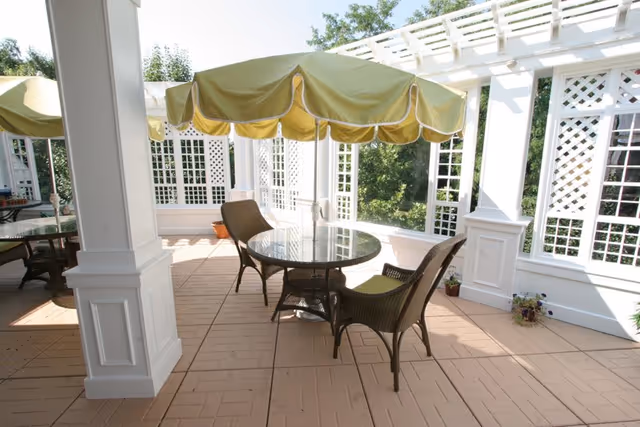 Covered outdoor patio with round glass-top tables, wicker chairs and yellow umbrellas beneath a white pergola.