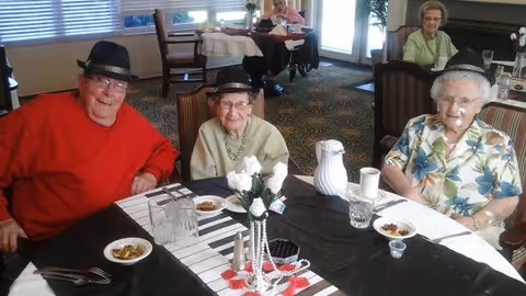 Three elderly residents wearing hats sit at a decorated dining table in a senior living facility dining room.