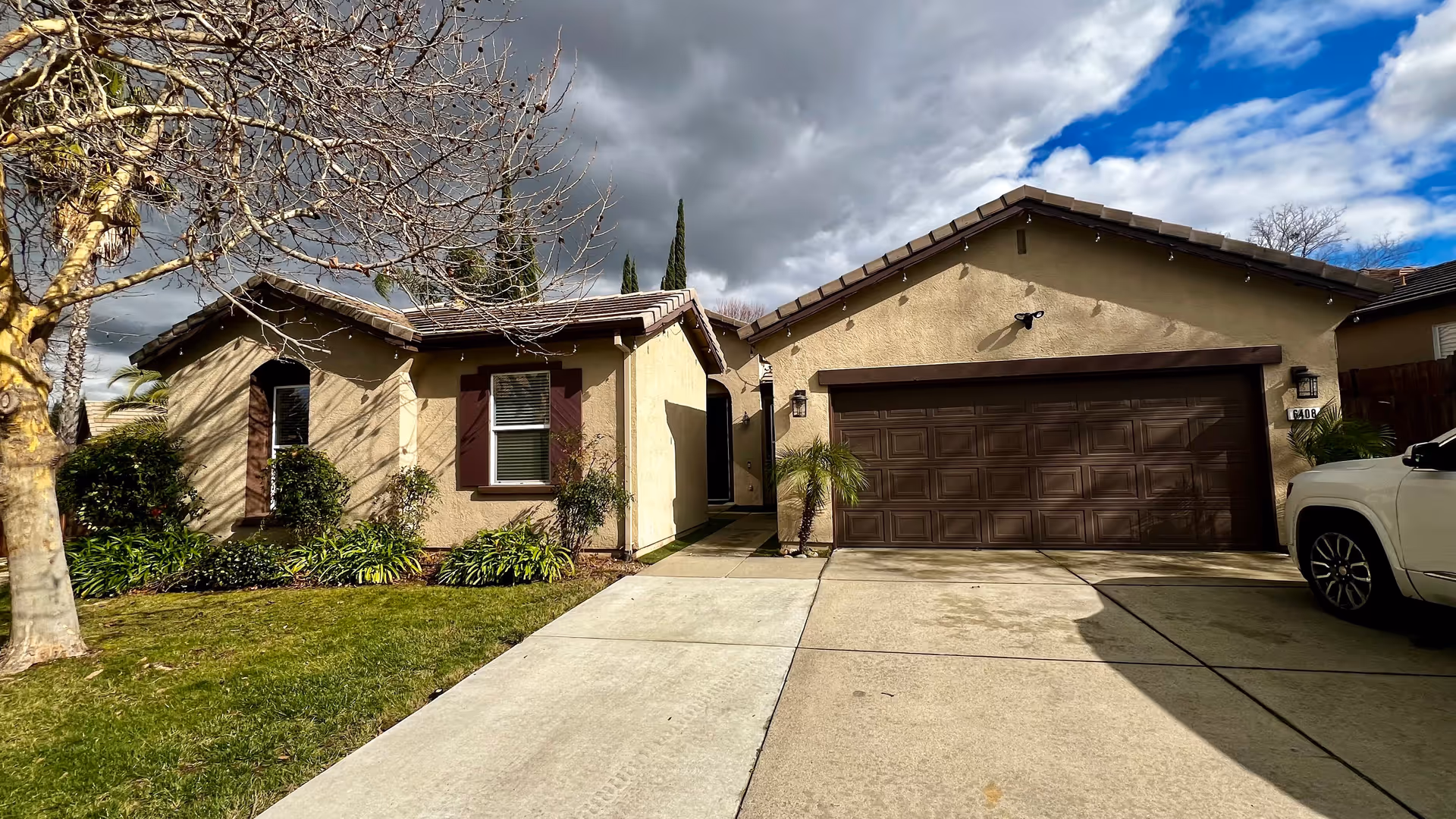 Front exterior view of a single-story residential building with beige stucco walls and a brown garage door. There is a driveway leading up to the garage, a small green lawn with plants and bushes, and a leafless tree on the left side. The sky is partly cloudy with patches of blue.