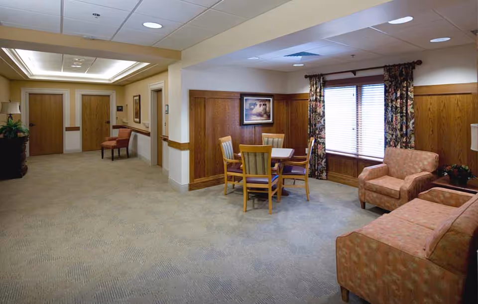 A cozy common area in a senior living facility featuring a small square table with four wooden chairs, a cushioned armchair, and a matching sofa. The room has wood paneling on the walls, a large window with floral curtains, framed artwork, and beige carpeting. There are two closed wooden doors and a hallway with additional seating visible in the background.